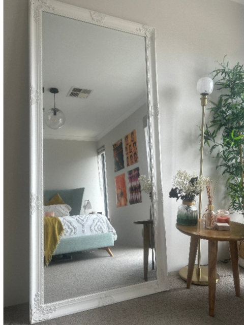 Large floor mirror with decorative frame in a room with a bed, table, and plants.
